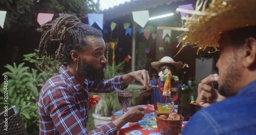 Group of happy black friends celebrating Festa Junina at an outdoor party with traditional food and drinks, wearing checkered shirts and straw hats under festive colorful flags in Brazil