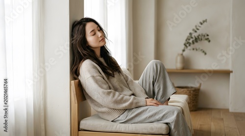 Young Asian woman sitting on the floor with eyes closed, enjoying a peaceful moment at home. Concept of mindfulness, relaxation, mental health, and serenity in a cozy, minimalist interior.