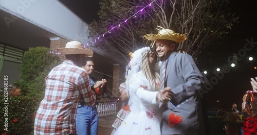 Traditional Brazilian Festa Junina celebration at night with people in caipira costumes dancing the quadrilha, featuring a bride and groom couple under festive lights and decorations