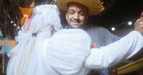 A happy couple dressed in traditional caipira wedding attire dances joyfully during a Brazilian Festa Junina celebration at night, surrounded by cheering guests and festive string lights