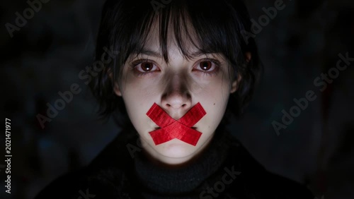 A close-up portrait of a young woman with red tape over her mouth forming an “X,” symbolizing silence, censorship, and emotional distress against a dark background.