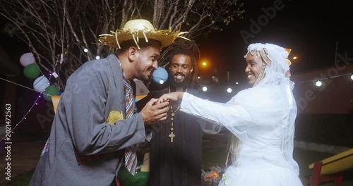 A joyful couple celebrates a traditional Brazilian Festa Junina wedding ceremony at night, with the groom in a straw hat kissing the bride's hand while a priest officiates the festive event under stri