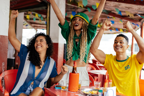 Brazilian friends enjoying drinks and watching football together at a casual bar, celebrating and socializing in a vibrant atmosphere.