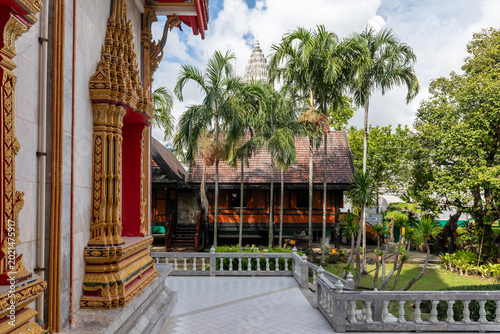 Photos Ornate golden carvings on a traditional Thai temple wall with red accents, overlooking lush palm trees and a wooden house under a bright blue sky with a distant stupa in Bangkok, Thailand