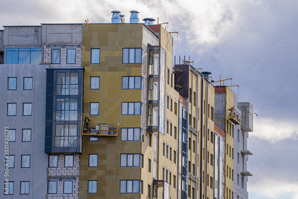 Fototapeta premium Building workers are applying thermal insulation panels and finishing materials to the exterior facade of a modern residential building, showcasing active construction work and urban development