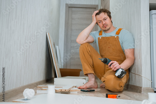 Man in overalls experiencing frustration and confusion while trying to assemble flat-pack furniture at home, holding a drill and looking at the disassembled parts