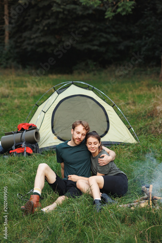 Young couple sitting near fire near tent in forest at summer