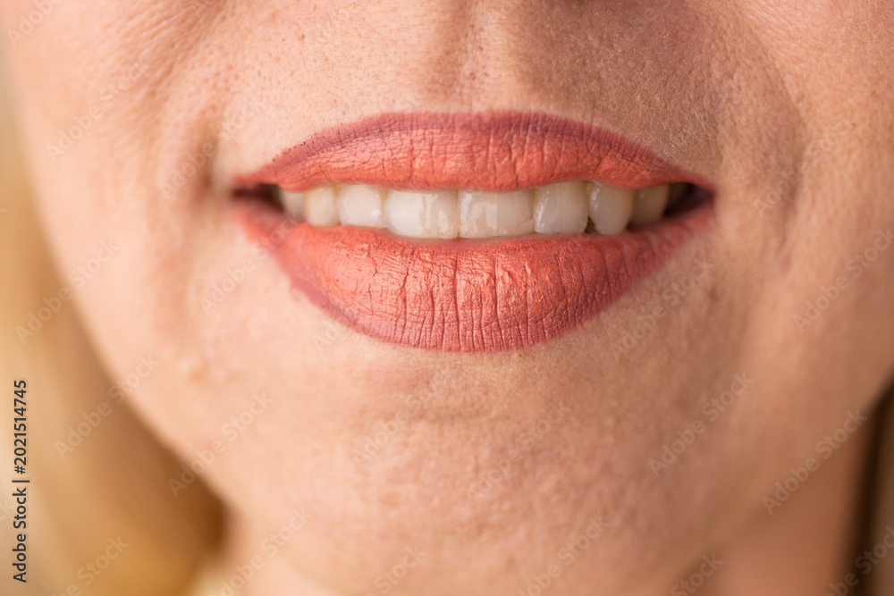 Fototapeta premium Caucasian woman smiling with coral lipstick on her lips. Macro shot of mouth showing white teeth. Beauty concept and professional makeup application for facial skin care presentation.