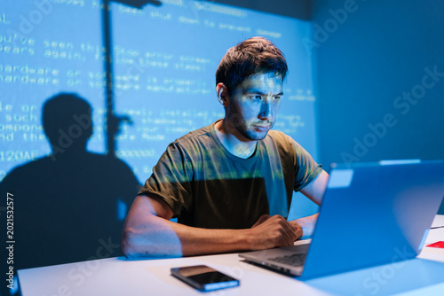Focused programmer concentrating on laptop, debugging code with projected lines on wall background, developing new software, working late in dark tech-driven environment.
