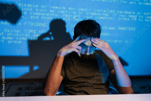 Stressed young male programmer facing challenging coding problem, feeling frustrated while working on laptop with lines of blue code projecting on wall behind him.