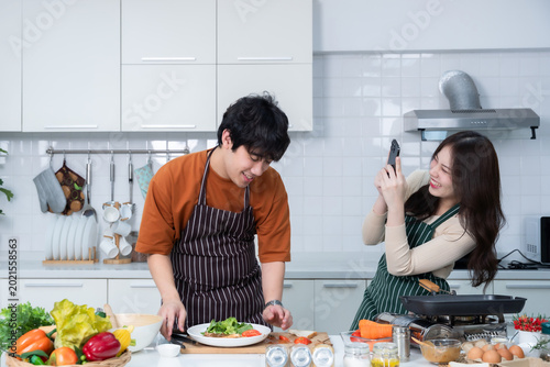 Happy of loving young asian Take a smartphone of having fun standing a cheerful preparing beef steak food and enjoy cook cooking with vegetables standing on a kitchen Condo life or home