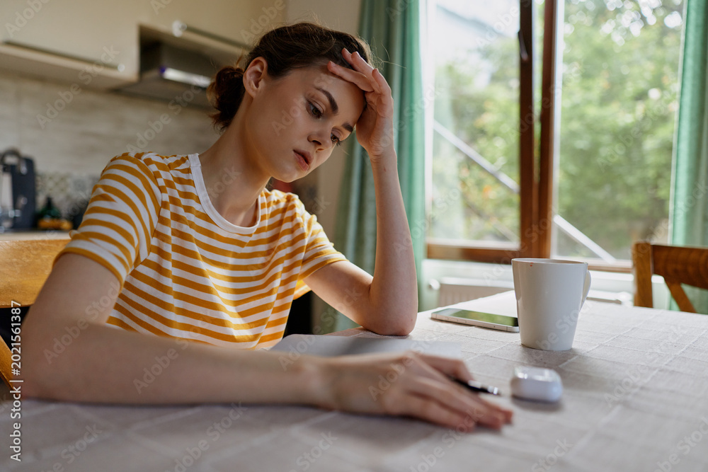 Naklejka premium Thoughtful woman indoors, resting hand on her forehead at home desk with notebook and mug, daytime light, striped shirt, reflective mood, casual lifestyle, focused moment