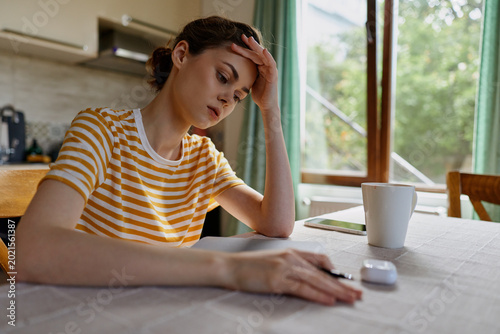Thoughtful woman indoors, resting hand on her forehead at home desk with notebook and mug, daytime light, striped shirt, reflective mood, casual lifestyle, focused moment