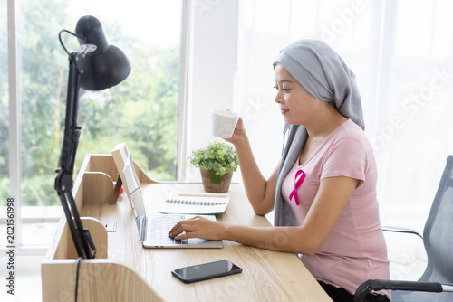 a asian women disease mammary cancer patient holding a coffee cup with pink ribbon wearing headscarf After treatment to chemotherapy with working business at laptop in office at home,medicine concept