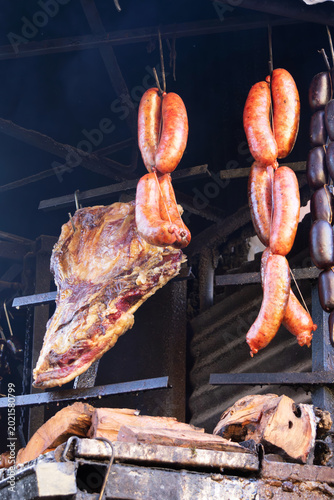 Assorted beef and sausages roast over an open wood fire. Warm, dramatic light highlights rich textures and smoky atmosphere. 