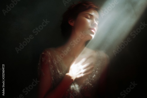 Woman with hand over heart illuminated by a soft beam of light for intimate portrait background and wellness poster design