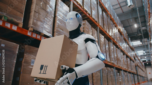 Fotografie White humanoid robot with glowing eyes carrying parcel through storage facility