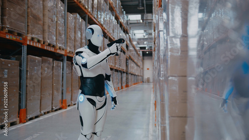 Intelligent humanoid robot pointing scanner at barcode while standing in warehouse aisle. Processing product information or tracking inventory updates during automated supply chain operation.