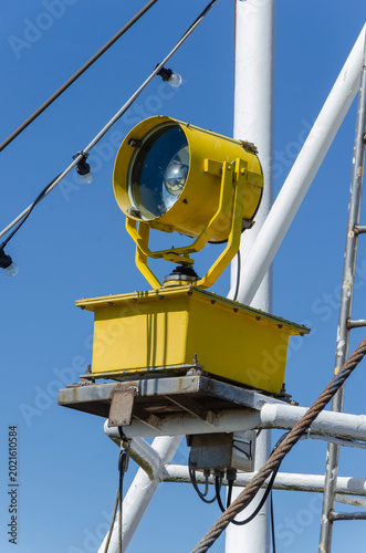 Yellow maritime searchlight mounted on a ship’s structure, surrounded by cables and metal fittings under a clear blue sky.