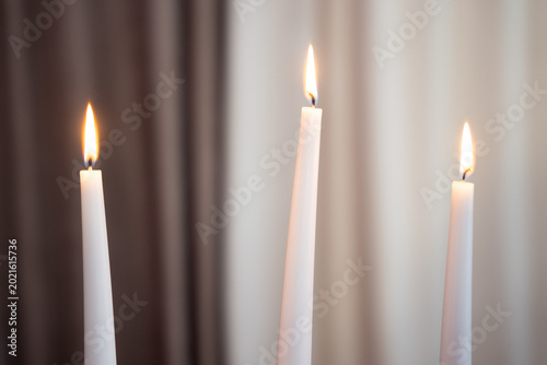 Photos Three burning white taper candles against a soft background