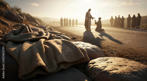 Worn woven cloak on stones with blurred biblical scene in background. Jesus healing a kneeling figure on a dusty road at sunset