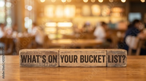 Wooden blocks spell out What's on your bucket list? sitting on a wooden table, with a blurred coffee shop in the background. Evoking thoughts of travel, life goals and adventure. 