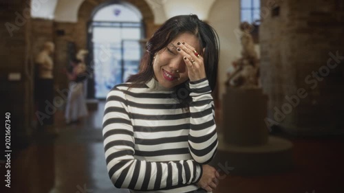 Young asian woman covers face with hand, smiling and tilting head, wearing black and white striped sweater beside sculptures on a pedestal in a building; playful shyness.