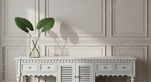 Elegant white wooden console table with intricate details and drawers, decorated with a glass vase holding two large green tropical leaves, set against a classic paneled wall in a tastefully decorated