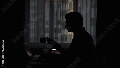 Fotografie Person silhouette sitting in dark room, working on laptop while drinking coffee,
