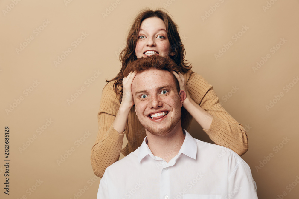 Fototapeta premium Happy young man and woman playfully pose together on beige background. The woman stands behind the man with hands on his head, both smiling joyfully. Concept of friendship and fun.