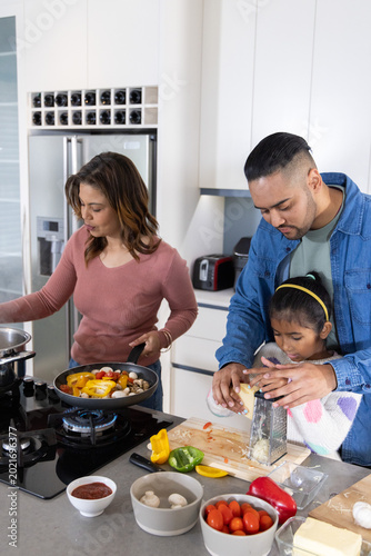 Family parents and child cooking vegetables on gas cooktop while grating cheese at kitchen island