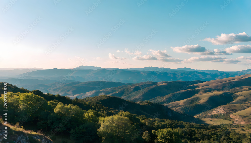 Obraz premium Rolling hills and mountains under a blue sky with scattered clouds and lush green trees