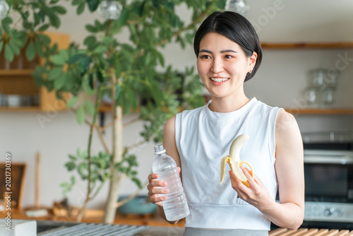 Quadro em tela A young Asian woman in sportswear eats a banana in the kitchen (training, energy replenishment, snack)