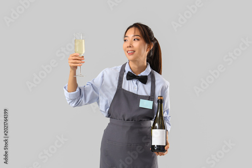 Young Asian smiling waitress with bottle and glass of wine on grey background