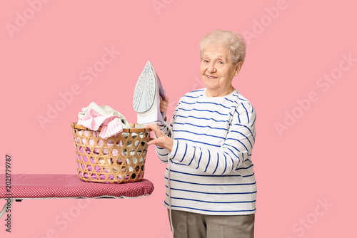 Happy senior woman with ironing board and basket of laundry on pink background