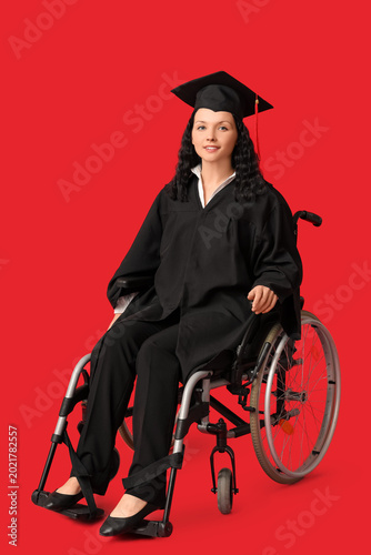 Beautiful female graduate student in wheelchair with graduation cap on red ba...
