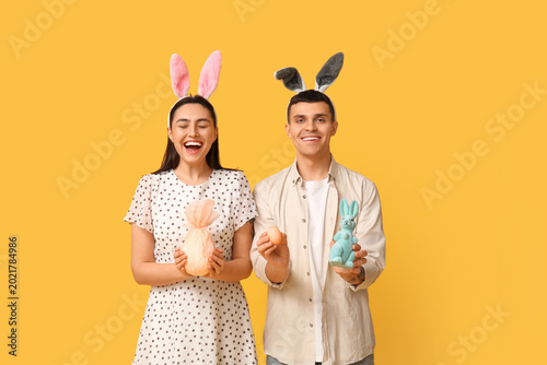 Happy young couple with Easter bunny ears, gift, toy rabbit and egg on yellow...