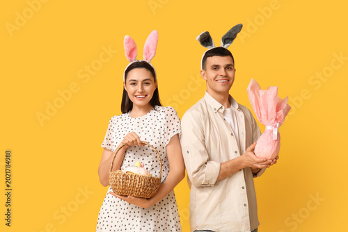 Happy young couple with Easter bunny ears, basket and gift on yellow background