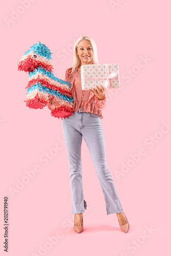 Happy mature woman with gift box and pinata on pink background