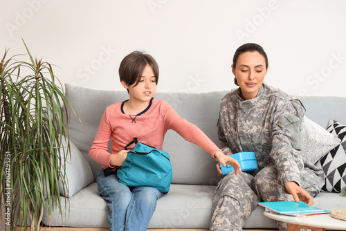 Military mother helping her little son to pack schoolbag in living room
