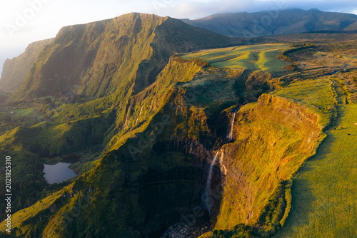 Aerial view of Poco da Ribeira do Ferreiro waterfalls cascading down lush green cliffs during golden hour in Fajazinha, Azores, Portugal.
