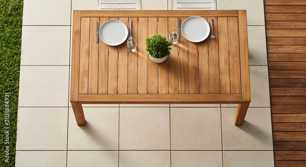 Fototapeta premium Overhead view of rectangular wooden dining table set for two on tiled patio, featuring two plates and small potted plant.