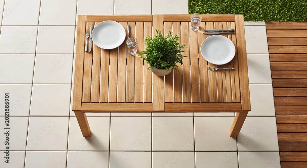 Fototapeta premium Top down view of small wooden outdoor table set for two, complete with plates, cutlery, and small potted plant, situated on tiled patio next to wooden deck and grass.