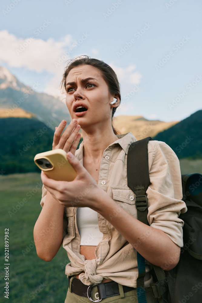 Fototapeta premium A woman outdoors with a backpack, holding a smartphone and reacting with surprise, standing in a mountain valley under a blue sky.