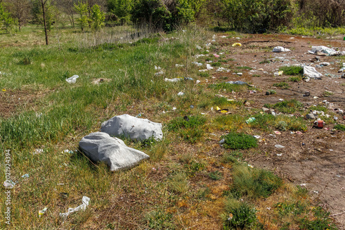 Household trash and plastic waste bags scattered across a green meadow and dirt ground during a sunny spring day