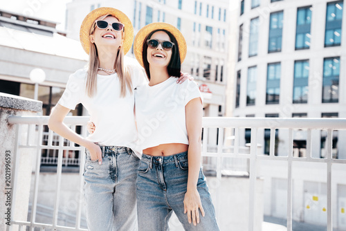 Two young beautiful smiling hipster female in trendy summer white t-shirt and jeans clothes. Carefree women posing in street. Positive models having fun outdoors. Cheerful and happy. In hat, eyewear