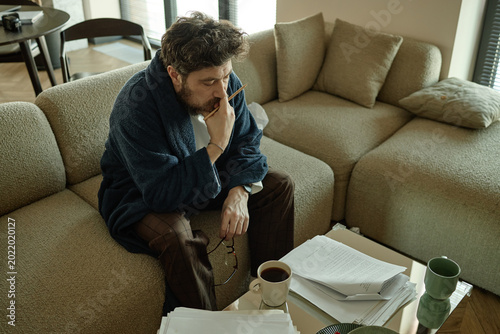 High angle view of middle aged Caucasian man sitting on sofa, reviewing paperwork and thinking with pencil, conveying stress and focus
