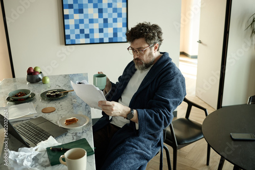 Middle aged Caucasian man wearing bathrobe reading new chapter of novel and drinking coffee beside laptop at home

