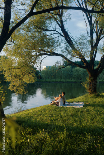 Couple enjoys a peaceful picnic by a serene pond under the shade of trees during golden hour
