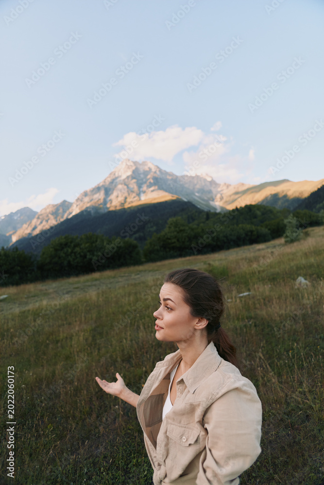 Fototapeta premium A young woman in casual beige jacket stands in a grassy field with mountains in the background under a clear sky. Lifestyle concept of nature exploration and outdoor relaxation.